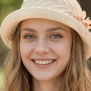 May include: A woman wearing a cream-colored sun hat with a floral accent. The hat has a woven texture and a wide brim. The woman has fair skin, green eyes, and is smiling. Her long, wavy hair is light brown.