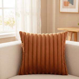 Peut inclure: Un coussin décoratif carré de couleur rouille avec une texture en velours côtelé. Le coussin est placé sur un canapé blanc. Le coussin a des rainures verticales et un aspect doux et moelleux. Décoration d'intérieur.
