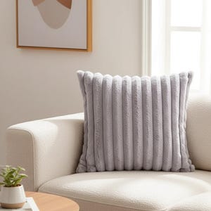 May include: A square, pale grey cushion with a ribbed, plush texture. The cushion is placed on a cream-coloured sofa. The design of the cushion adds a touch of elegance and comfort to the living space.