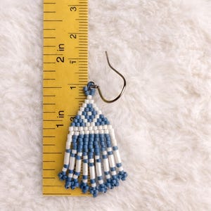 May include: Handmade beaded earrings with a blue and white geometric design. The earrings are approximately 1.5 inches long, featuring a triangular top and dangling fringe. The earrings are displayed on a white fluffy surface.