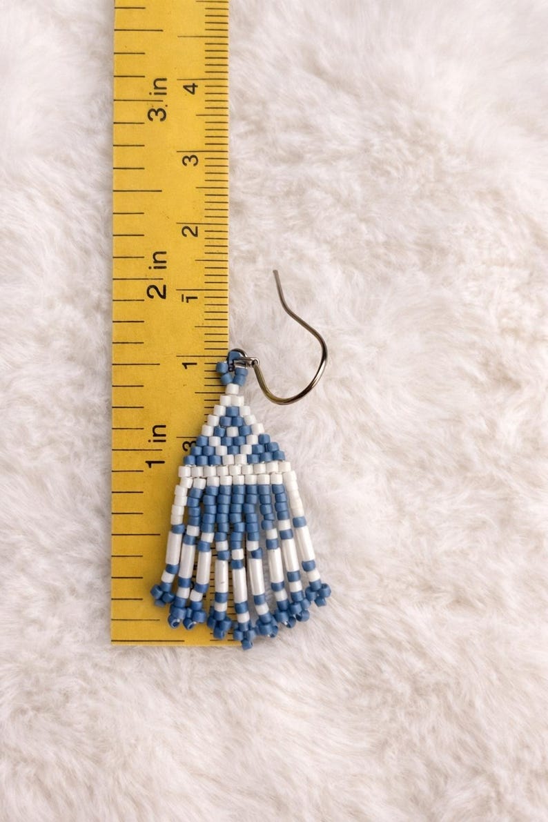 May include: Handmade beaded earrings with a blue and white geometric design. The earrings are approximately 1.5 inches long, featuring a triangular top and dangling fringe. The earrings are displayed on a white fluffy surface.