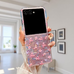 Peut inclure: Une coque de téléphone transparente avec un motif floral. La coque présente des fleurs roses et de petits papillons sur un fond nacré. La coque est ouverte, révélant un écran noir et un appareil photo.