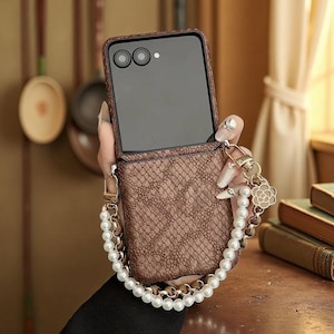 Könnte beinhalten: Braune, strukturierte Handyhülle mit einer Perlen-Handschlaufe und goldfarbener Hardware. Die Hülle ist geöffnet und zeigt den Bildschirm und die Kameralinsen des Telefons. Ein dekorativer Anhänger mit Blumenmuster ist befestigt.