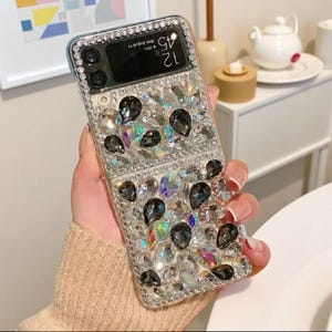 Puede incluir: Una funda transparente para teléfono incrustada con pedrería y gemas en forma de lágrima de color gris oscuro. La funda tiene un marco plateado y un área de cámara negra. El teléfono muestra la hora 12:45 el miércoles 11 de agosto.