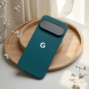 May include: Teal phone case with a large camera cutout and a white "G" logo. The case is on a wooden tray. The case is designed to protect the phone's camera. The background is light-coloured.