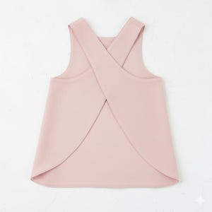 Könnte beinhalten: Ein ärmelloses, blassrosa Oberteil mit einem einzigartigen gekreuzten Rückendesign. Das Oberteil hat einen runden Ausschnitt und eine fließende A-Linien-Silhouette. Die Träger kreuzen sich am Rücken und erzeugen ein interessantes visuelles Detail. Der Stoff scheint ein weiches, leichtes Material zu sein.