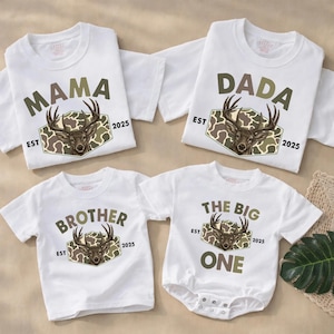 May include: White t-shirts and a onesie with matching deer and camouflage designs. The shirts read "MAMA", "DADA", "BROTHER", and "THE BIG ONE". Each shirt has "EST 2025" printed below the deer graphic.