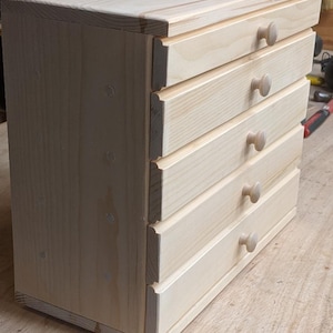 May include: A small, unfinished wooden chest of drawers with four drawers. The chest is made of light-colored wood and features round wooden knobs. The drawers are evenly spaced and the overall design is simple and functional.