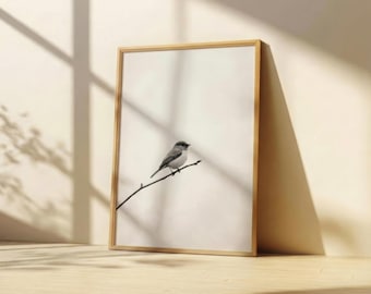 May include: A framed print featuring a small bird perched on a slender branch. The bird is depicted in shades of gray and white against a neutral background. The frame is made of light-colored wood, and the artwork is leaning against a wall.