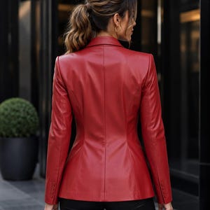 May include: A red leather blazer with a tailored fit. The blazer features a classic notched lapel, buttoned cuffs, and a structured silhouette. The back view shows the blazer's seams and design details. The model is wearing black pants.