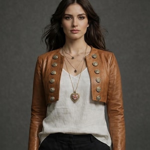 Puede incluir: Chaqueta bolero de cuero marrón con botones dorados. La chaqueta se lleva sobre una camiseta blanca y combinada con pantalones negros. Un collar dorado en capas con un colgante de corazón completa el look. Chaqueta elegante.