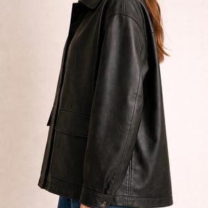 May include: Black faux leather jacket with a collared neckline, two front pockets, and buttoned cuffs. The jacket has a classic, slightly oversized fit, suitable for various outfits.