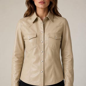 May include: A beige long-sleeve button-down shirt made of a smooth material with a pointed collar, two chest pockets with flaps, and silver-toned snap buttons down the front placket and on the cuffs, paired with black pants.