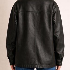 May include: A black leather jacket with a collared neckline and buttoned cuffs. The jacket has a classic design with a button-down front and a tailored fit. The model is wearing blue jeans.