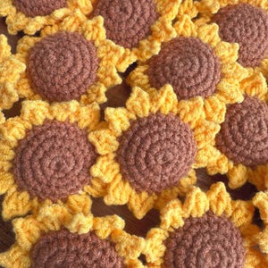 May include: Close-up of several handmade crochet sunflowers. Each flower has a brown centre and yellow petals. The sunflowers are arranged closely together, showcasing the texture and detail of the yarn.