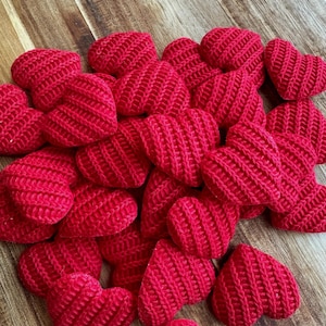 May include: A cluster of small, knitted red hearts on a wooden surface. Each heart features a textured, ribbed design. The hearts are a bright red colour, creating a visually appealing arrangement.