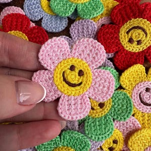 May include: A selection of colourful, crocheted flower appliques. Each flower has a smiley face in the centre and comes in shades of pink, red, green, yellow, and purple. The petals have a textured appearance, giving a cheerful, handmade feel.