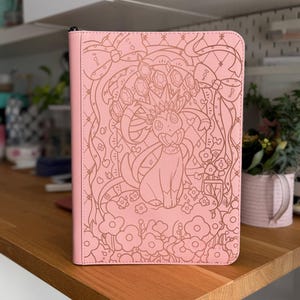 Peut inclure: Un classeur rectangulaire rose clair orné d'une illustration en traits gravés représentant un personnage Sylveon entouré de fleurs, de rubans et d'une arche de style vitrail. La couverture a une finition mate lisse avec une tirette noire.