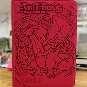 Puede incluir: Una carpeta de tarjetas de color rosa brillante con una ilustración de líneas de color rojo oscuro de la criatura Charizard de la franquicia Pokemon. La palabra Evolutions está impresa en la parte superior y la marca vaultx está grabada.