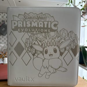 Puede incluir: Una carpeta para tarjetas coleccionables VaultX de color blanco con un gráfico en tono dorado del Pokemon Eevee rodeado de cristales. La portada muestra el texto Scarlet & Violet Prismatic Evolutions y un patrón de diamantes. Diseño estructurado.