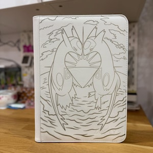 Peut inclure: Un classeur rectangulaire blanc orné d'une illustration au trait doré représentant deux créatures Latias et Latios se faisant face au-dessus d'un soleil stylisé et de vagues. Le motif est centré sur la couverture avant avec un style épuré.