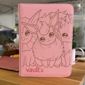 Peut inclure: Un classeur à cartes à collectionner rectangulaire rose clair avec une surface texturée. La couverture présente une illustration au trait rose foncé de trois créatures, dont Pyroli au centre. La marque vaultx est imprimée en rose foncé.