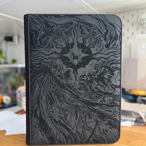 Peut inclure: Un classeur de cartes à collectionner rectangulaire noir avec une surface texturée présentant un motif graphique abstrait gris foncé composé de tourbillons et d'un emblème central en forme d'étoile. La marque Vault X est embossée en bas à gauche.