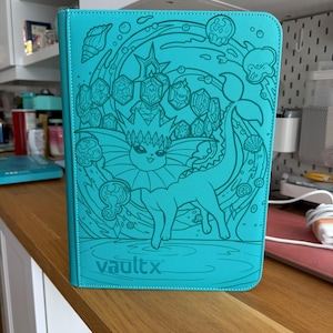 Peut inclure: Un classeur de cartes turquoise vif orné d'une illustration au trait bleu sarcelle foncé représentant un personnage Vaporeon entouré de bulles d'eau, de coquillages et de cristaux. Le logo VaultX est embossé au centre en bas de la couverture.