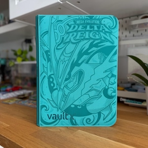 Peut inclure: Un classeur de cartes à collectionner de couleur sarcelle avec un motif en relief représentant une créature et le texte "MEGA EVOLUTION DELTA REIGN" en haut. Le logo "vaultx" est visible en bas à gauche. Le classeur a une surface texturée.