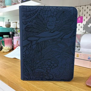 Peut inclure: Un classeur rectangulaire bleu foncé orné d'une illustration détaillée en lignes noires représentant une créature fantastique reposant sur une grande feuille, entourée d'un feuillage tropical dense, de fougères et de plantes diverses.