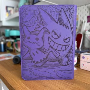 Peut inclure: Un classeur rectangulaire violet présentant une illustration au trait violet foncé des personnages Gengar et Mimikyu sur la couverture avant. Le design comprend des lignes complexes détaillant les créatures et un motif de fond texturé.