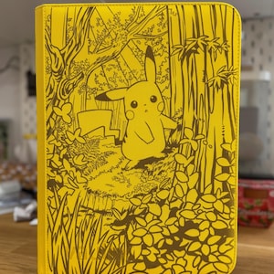 Puede incluir: Carpeta amarilla con un diseño de Pikachu en un entorno forestal. La ilustración marrón muestra a Pikachu entre árboles y follaje. La carpeta rectangular probablemente está hecha de un material duradero, ideal para organizar papeles o tarjetas.