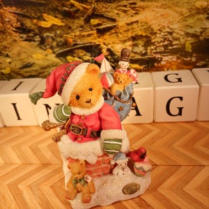 Op de afbeelding: Een kerstbeeldje met een teddybeer in een rode jas en hoed, bovenop een schoorsteen. Het beeldje bevat een kleine teddybeer, een sneeuwpop en een tinnen soldaat. De basis is versierd met sneeuw en een klein bordje.