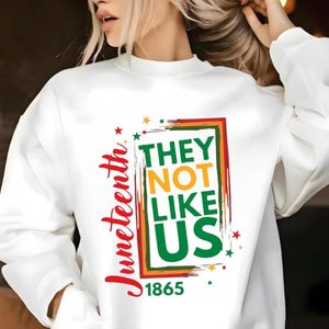 Może przedstawiać: Biała bluza z napisem "Juneteenth" w czerwonym skrypcie. Projekt zawiera frazę "THEY NOT LIKE US" w zielonej, pomarańczowej i czerwonej ramce, z rokiem "1865" poniżej. Gwiazdy są rozrzucone wok&oacute;ł projektu.