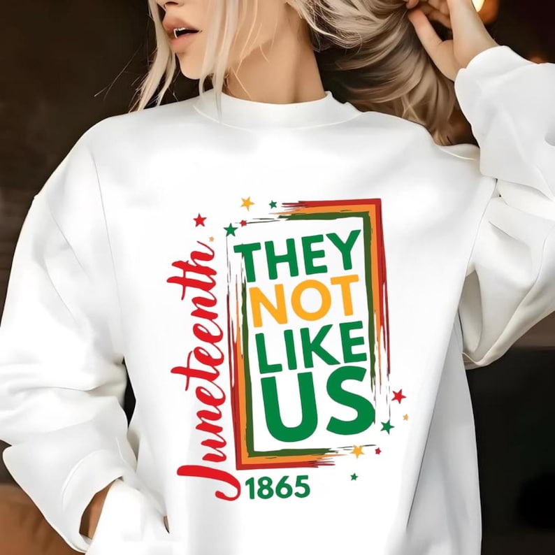 Może przedstawiać: Biała bluza z napisem "Juneteenth" w czerwonym skrypcie. Projekt zawiera frazę "THEY NOT LIKE US" w zielonej, pomarańczowej i czerwonej ramce, z rokiem "1865" poniżej. Gwiazdy są rozrzucone wok&oacute;ł projektu.