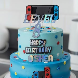 May include: A two-tiered birthday cake with blue icing, decorated with star-shaped sprinkles and a video game controller theme. The top tier has a "Level 8" cake topper. The cake says "Happy Birthday O'Shea".