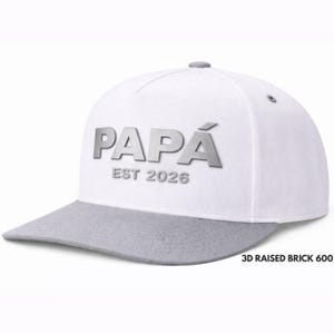 May include: A white and gray baseball cap with the word "PAPÁ" in silver, and the text "EST 2026". The cap has a flat brim and a gray top button. The hat is a stylish accessory.