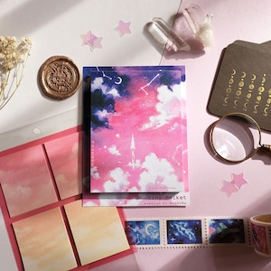 May include: A set of pink and blue watercolor sticky notes with a galaxy theme. The notes feature clouds, stars, and a rocket ship. The set also includes a roll of galaxy-themed washi tape.