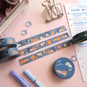 May include: Washi tapes with cat designs in blue, purple, and gold. The tapes are displayed with a clear storage box, a planner, and stationery items on a pink surface. The cat illustrations show various poses. The scene is well-lit.