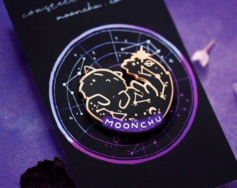 May include: Gold and black enamel pin featuring a curled up cat with stars and constellations on its fur. The pin is on a black card with the text "MOONCHU" in purple.