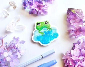 May include: A green frog with big eyes sits on a blue and white cloud with white stars. The sticker is on a white background with purple flowers and clear crystals.