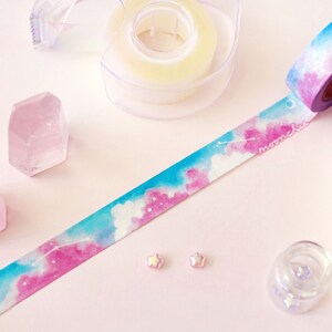 Op de afbeelding: Een rol washi tape met een aquarelontwerp van roze en blauwe wolken en een halve maan. De tape ligt op een roze oppervlak met twee roze kristallen en twee kleine roze sterren.