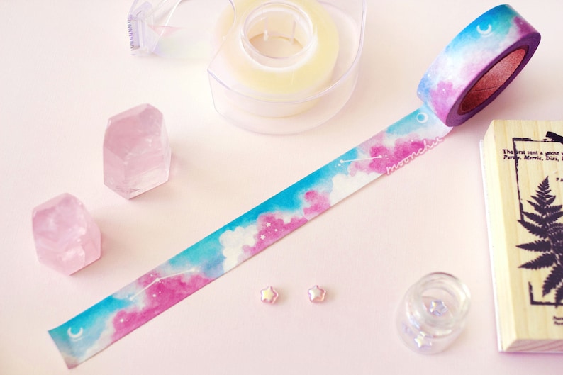 Op de afbeelding: Een rol washi tape met een aquarelontwerp van roze en blauwe wolken en een halve maan. De tape ligt op een roze oppervlak met twee roze kristallen en twee kleine roze sterren.