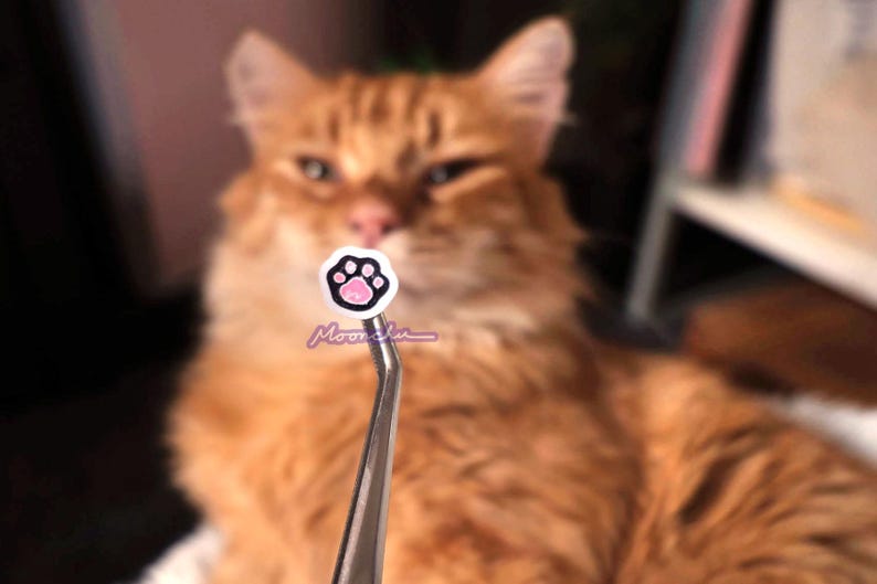 Puede incluir: Primer plano de una peque&ntilde;a pegatina de huella de pata rosa en unas pinzas de plata. La pegatina est&aacute; enfocada, mientras que las pinzas y un gato naranja borroso est&aacute;n desenfocados en el fondo.
