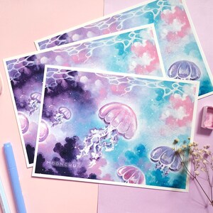 Puede incluir: Un conjunto de tres impresiones de acuarela con un fondo de tipo galaxia en tonos morados, rosas y azules. Las impresiones representan medusas con cuerpos transl&uacute;cidos y detalles brillantes. Las impresiones est&aacute;n firmadas "MOONCHU" en la esquina inferior derecha.