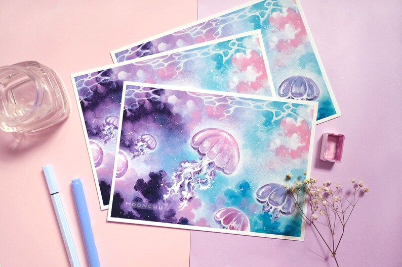 Puede incluir: Un conjunto de tres impresiones de acuarela con un fondo de tipo galaxia en tonos morados, rosas y azules. Las impresiones representan medusas con cuerpos transl&uacute;cidos y detalles brillantes. Las impresiones est&aacute;n firmadas "MOONCHU" en la esquina inferior derecha.