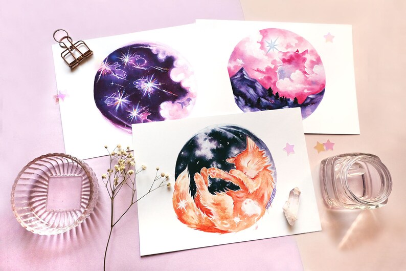 May include: Three watercolor paintings on white paper with a pink background. The first painting depicts a purple and pink night sky with white clouds and shooting stars. The second painting shows a pink and purple sunset with a mountain range in the background. The third painting features a sleeping orange cat with a starry night sky in the background.