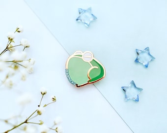 May include: A green frog enamel pin with a gold outline and the word "MOENCHU" written on the side. The pin is on a light blue background with white flowers and blue star-shaped decorations.