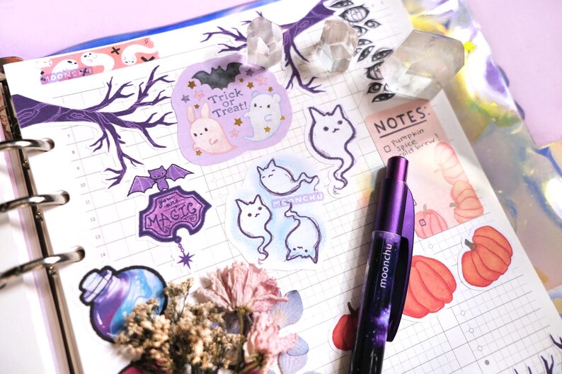 Puede incluir: Un planificador morado con una variedad de pegatinas, incluyendo una botella de poci&oacute;n morada, un cartel que dice "You're Magic", una rama de &aacute;rbol, un fantasma y un gato. El planificador tambi&eacute;n tiene una nota que dice "Pumpkin Spice Did You Feel It?" y una pluma morada.