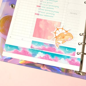 Op de afbeelding: Een witte planner met een transparante plastic hoes met een holografisch ontwerp. De planner heeft een ringband en een pagina met een roze aquarel achtergrond. Een sticker van een walvis met een ster op zijn rug is op de pagina. De pagina heeft ook een decoratieve washi tape met een roze en blauwe hemel en sterren ontwerp.